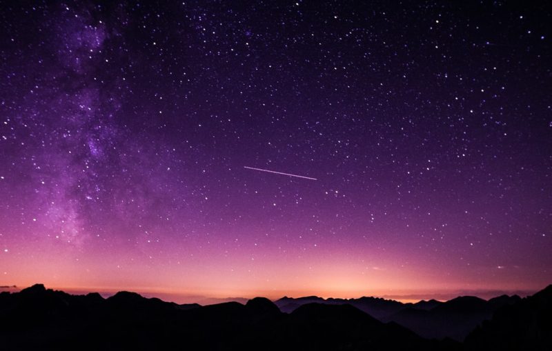 a purple-pink starry sky. a small comet streaks across. the foreground is a dark silhouette of mountains.