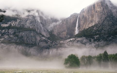 a closeup of a mountain range, partially covered in fog. some trees can be seen on the ground below.