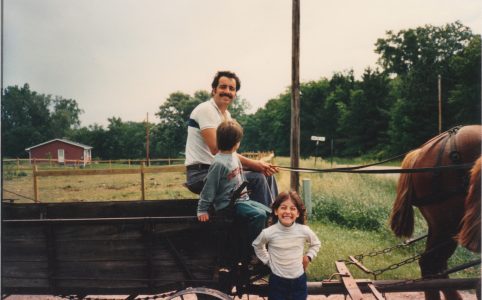 a dark-haired man with a mustache in a short sleeve white shirt is sitting on an old wooden manure spreader, holding the reins of a pair of auburn Belgian workhorses. a young child sits next to him turned away from the camera. another child faces the photographer with a big smile.