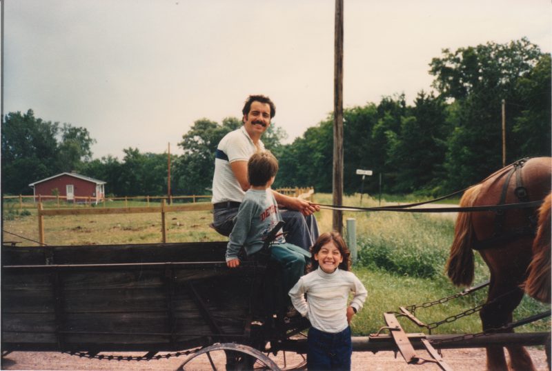 a dark-haired man with a mustache in a short sleeve white shirt is sitting on an old wooden manure spreader, holding the reins of a pair of auburn Belgian workhorses. a young child sits next to him turned away from the camera. another child faces the photographer with a big smile.