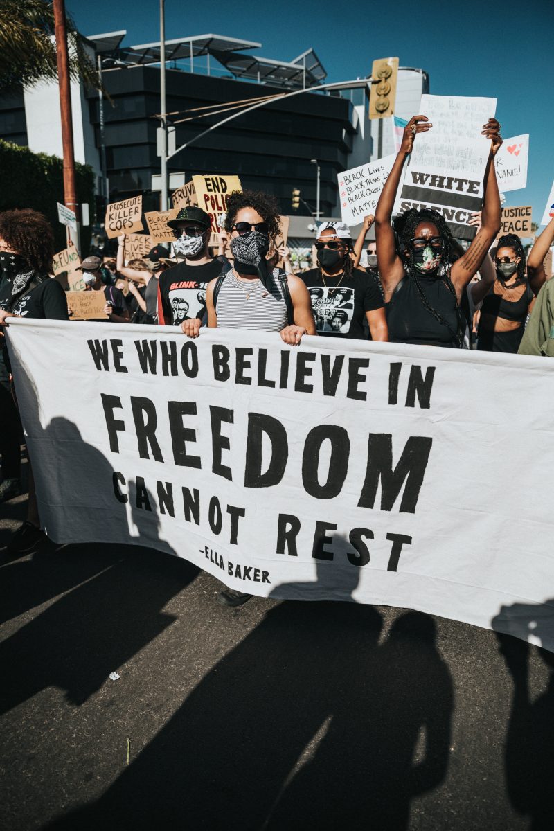 people marching with many signs, including a large sign held by several people, white with black letters that reads "we who believe in freedom cannot rest" by Ella Baker