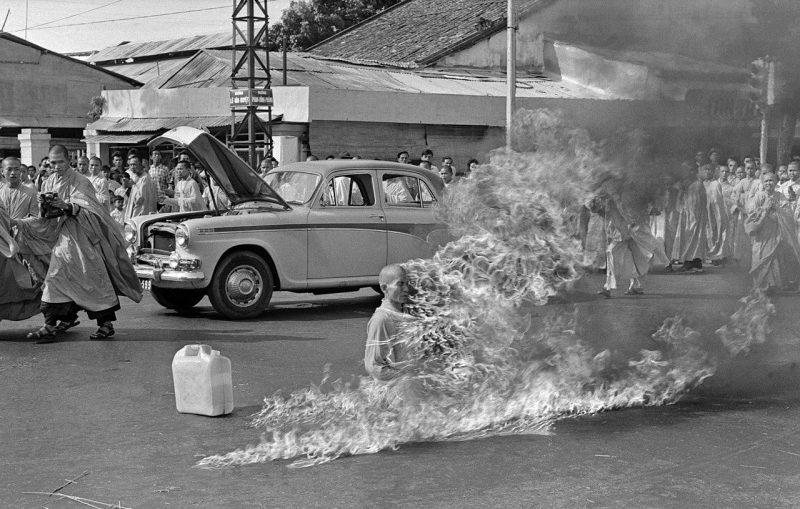 a black and white photo of a street with people in robes and a car in front of a cluster of buildings. the focal point is a monk who is actively on fire. this is the famous photo of Quảng Đức during his self-immolation
