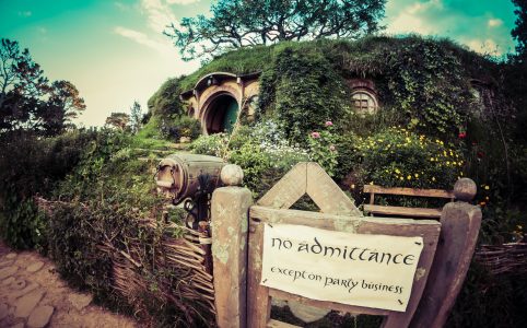 a scene of a hobbit house surrounded by flowering plants and a wicker fence, with a sign reading 'no admittance except on party business' nailed to the gate