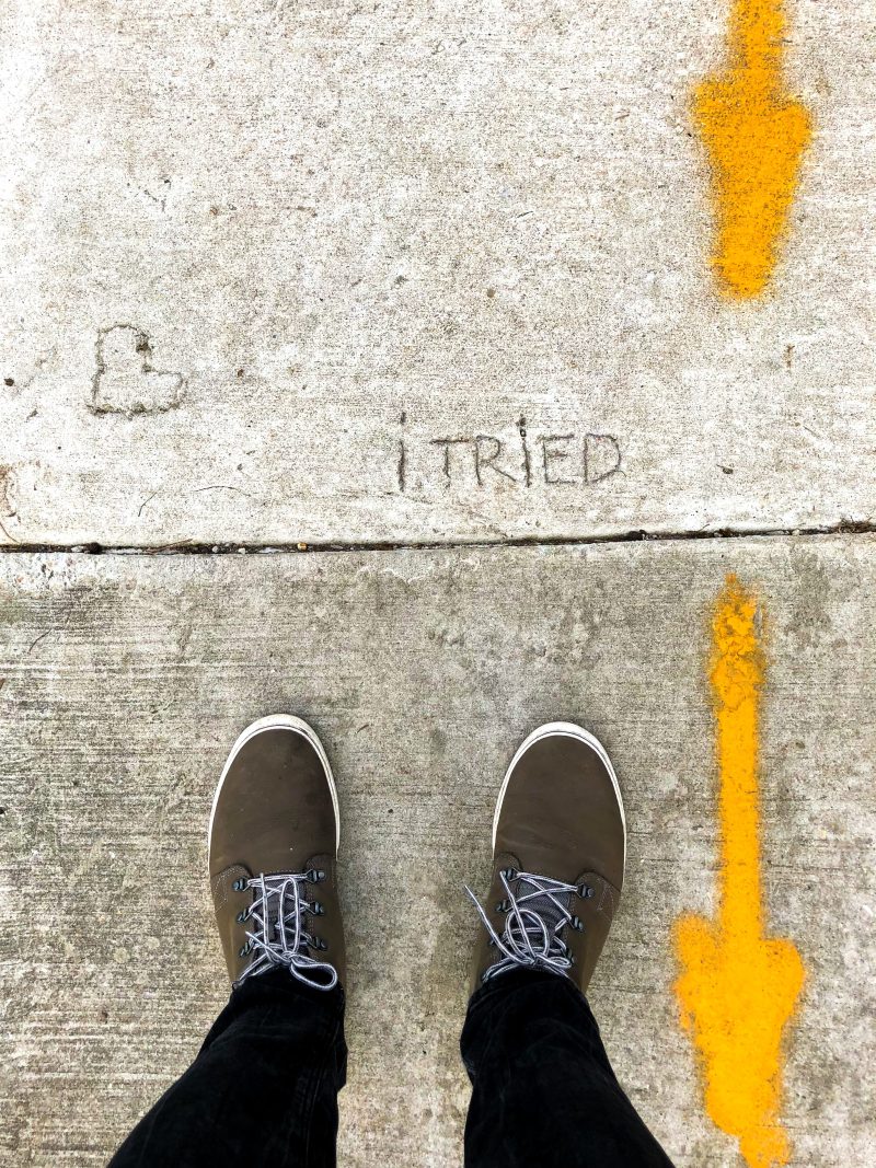 a POV looking downward at two feet on a sidewalk. the pavement is painted on the right with yellow arrows pointing downward, and scratched into the pavement above the feet is the phrase 'i tried'