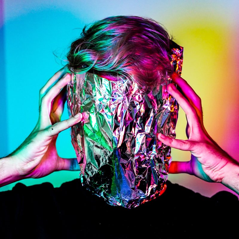 a person with aluminum foil obscuring their face, with hands on both sides of their face, in multicolored light