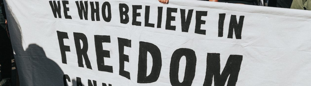 people marching with many signs, including a large sign held by several people, white with black letters that reads "we who believe in freedom cannot rest" by Ella Baker