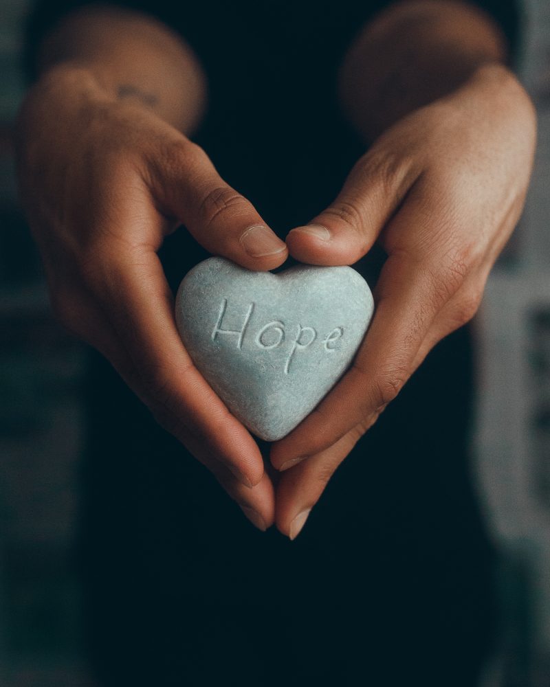 two brown hands holding a heart-shaped stone carved with the word 'Hope'