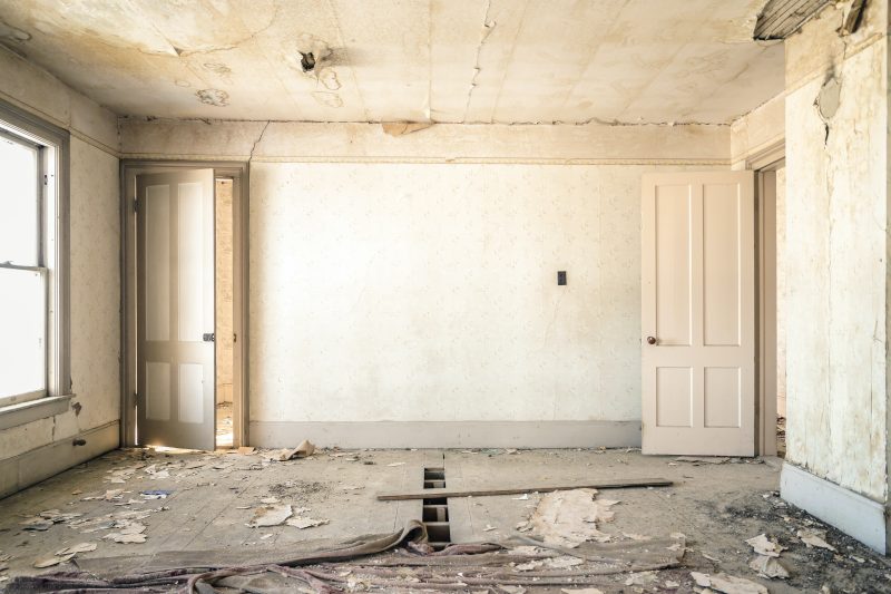 interior photo of a room that is old and beginning to fall apart. the paint and wallpaper are peeling and there are broken wooden slats in the floor.