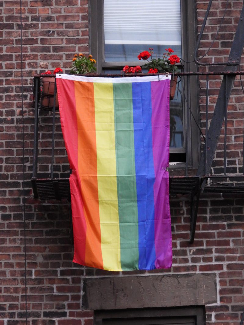 a rainbow flag hangs from the metal balcony of a brick building