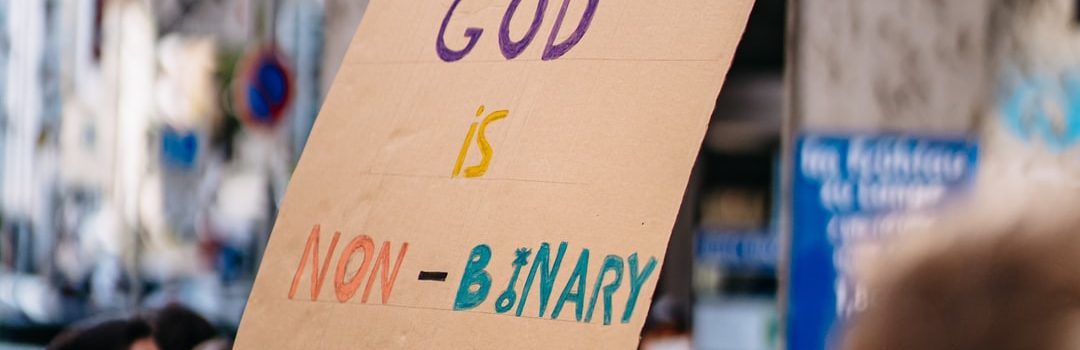 in a crowd at a peaceful protest gathering, a person in a white face mask and dark hair with glasses holds a sign that reads 'GOD IS NON-BINARY'