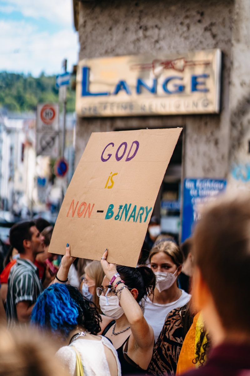 in a crowd at a peaceful protest gathering, a person in a white face mask and dark hair with glasses holds a sign that reads 'GOD IS NON-BINARY'