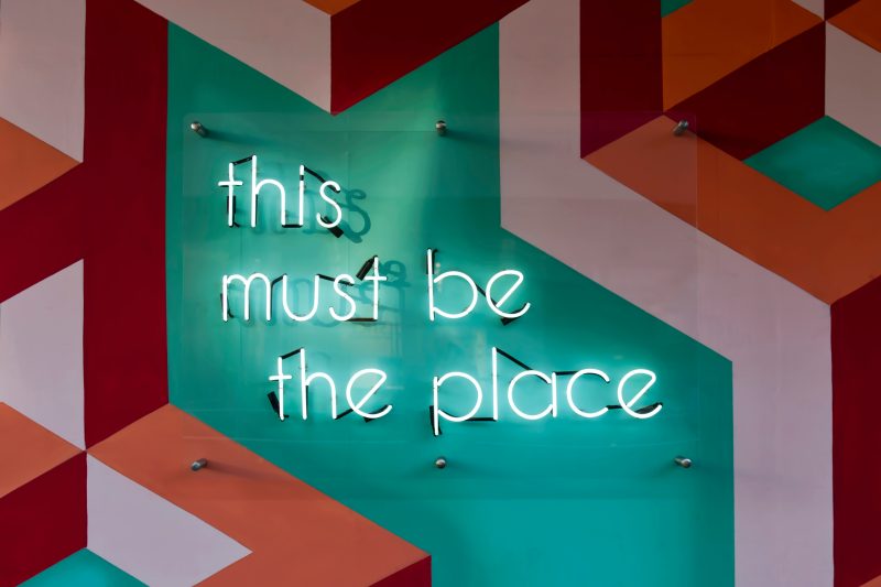 an orange, white, red, and turquoise wall with a neon sign with the words 'this must be the place'