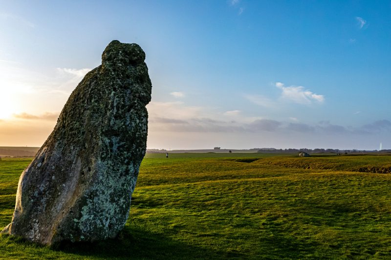 a photo of sunlight off-camera to the left, with a leaning ancient stone in front of the light. there is a grassy field and the sky is blue with some clouds.