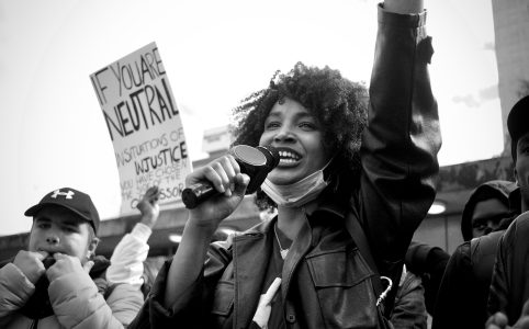 a black and white photo of a protest. the person in the forefront has dark skin and a microphone. there is a sign on the left that reads "If you are neutral in situations of injustice, you have chosen the side of the oppressor."
