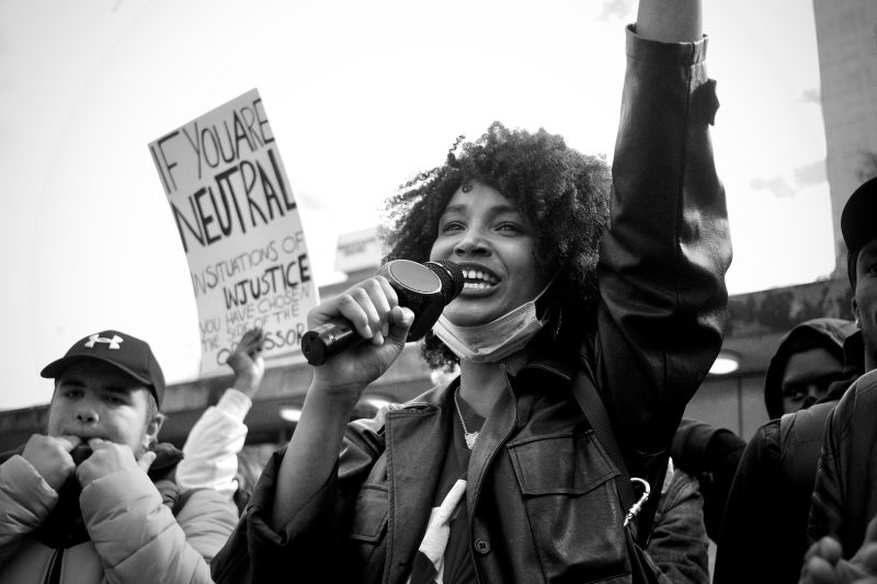 a black and white photo of a protest. the person in the forefront has dark skin and a microphone. there is a sign on the left that reads "If you are neutral in situations of injustice, you have chosen the side of the oppressor."