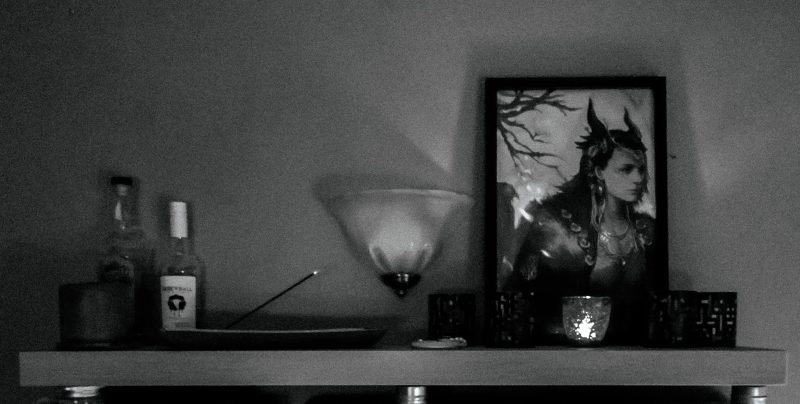a black and white photo of my Loki shrine, a shelf with candles, incense, and a large framed likeness of the god Loki