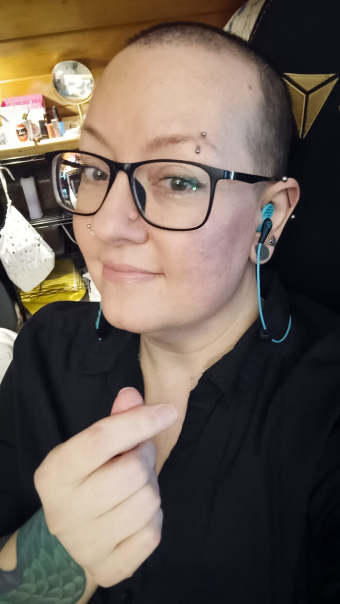 selfie of a pale person with dark hair, black glasses, piercings, and a tattoo, wearing a black shirt and turquoise earbuds.